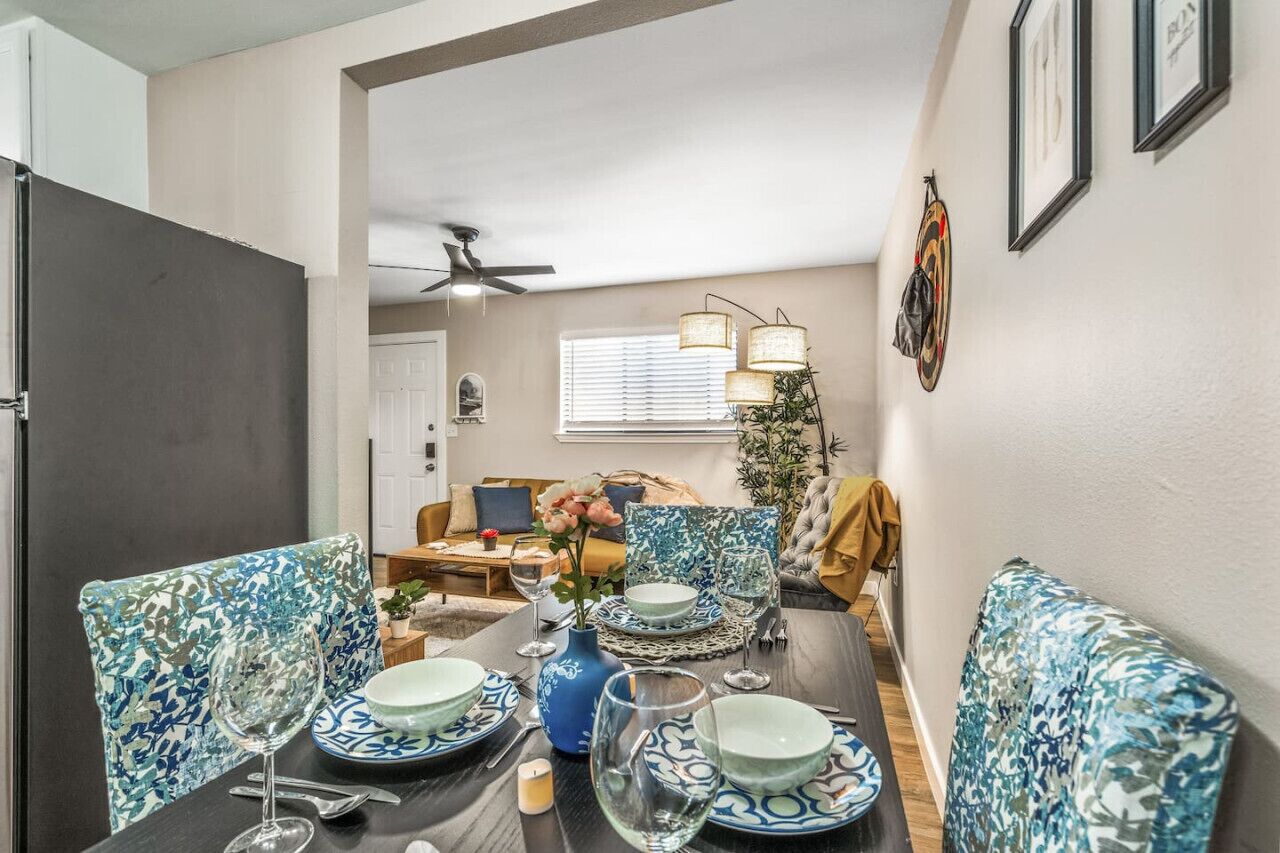 A dining area with a table set for four, chairs with blue floral upholstery, a vase with flowers, and a ceiling fan.