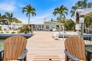A wooden dock leading to a house with a pool, surrounded by palm trees and a clear sky.