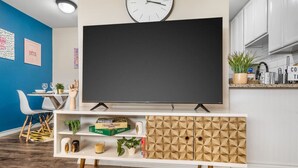 A modern living room with a flat-screen TV, a blue accent wall, a wooden cabinet with geometric patterns, and a small dining area with a white chair and table. - Where It's All Happening: 1BD in the Mid of Action (Arlington)