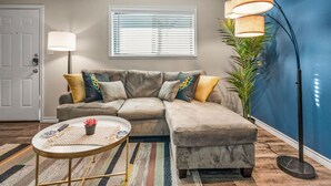 A living room with a sectional sofa, a round coffee table, a floor lamp, a potted plant, and a window with blinds. - Where It's All Happening: 1BD in the Mid of Action (Arlington)