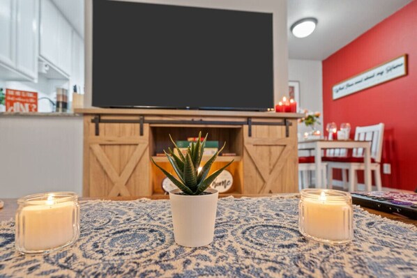 A cozy living room with a flat-screen TV mounted above a wooden fireplace, a potted plant, and two lit candles on a patterned rug.