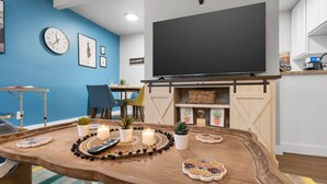 A modern living room with a large TV, a wooden coffee table with decorative items, a blue accent wall, and a cozy seating area. - Sapphire 1BD: AT&T Walkable, Calming & Joyful ! (Arlington)