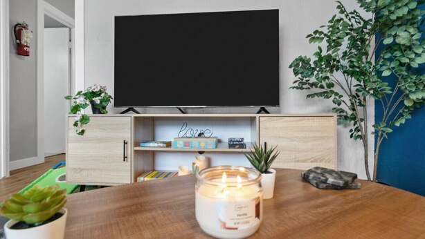 A living room with a flat-screen TV mounted on the wall, a wooden table with a lit candle, and a potted plant. - Blissful 1BD:AT&T Walkable, Modern & Ground-Level! (Arlington)