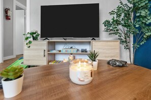 Un salon avec un téléviseur à écran plat fixé au mur, une table en bois sur laquelle est posée une bougie allumée et une plante en pot.