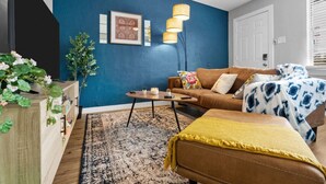 A living room with a blue accent wall, a brown sofa, a yellow throw, and a coffee table with decorative items. - Blissful 1BD:AT&T Walkable, Modern & Ground-Level! (Arlington)