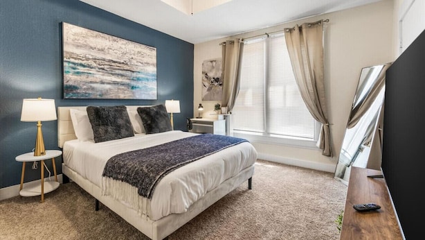 A bedroom with a bed, bedside lamps, a large painting, a window with curtains, and a TV on a wooden stand. - Uplifting 2BD: Centered, Inspiring & Heartwarming! (Fort Worth)