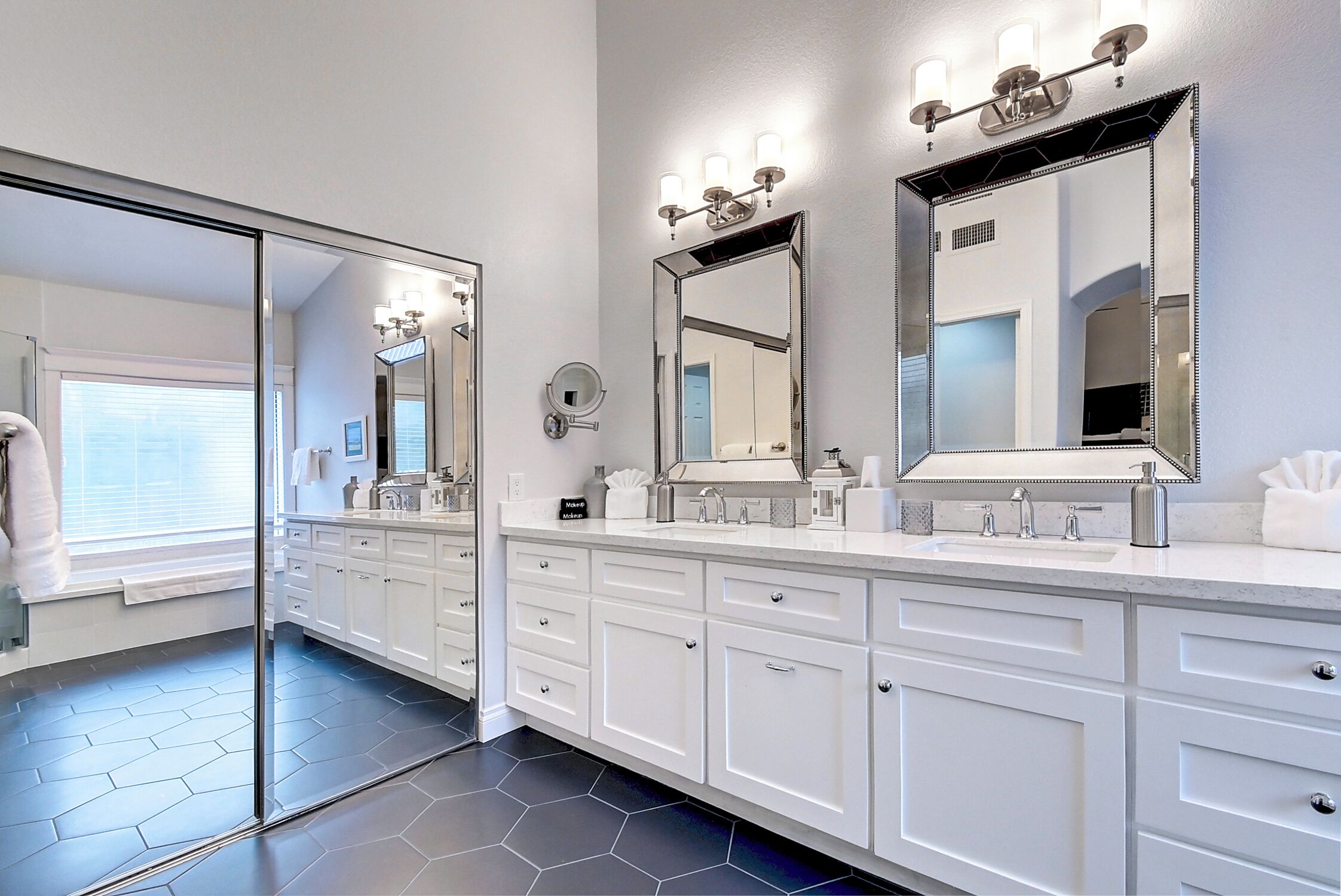 A modern bathroom with a large mirror, white cabinetry, and a hexagonal tile floor.