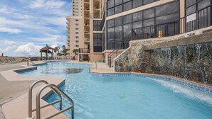 A swimming pool with a stone waterfall feature, surrounded by a multi-story building with balconies. - GULF-FRONT CONDO, POOLS, SPLASH PAD, LOTS OF AMENITIES! (Orange Beach)