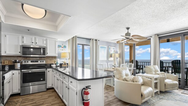 A modern kitchen with white cabinetry, stainless steel appliances, and a large island. There is a dining area with white chairs and a table, and a balcony with a view of the sky. - GULF-FRONT CONDO, POOLS, SPLASH PAD, LOTS OF AMENITIES! (Orange Beach)