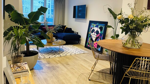 A modern living room with a blue sofa, a coffee table, and a large window. There is a painting of a panda on the wall and a vase with flowers on a side table. - Modern apartment in the heart of London.  (London)