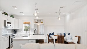 Une cuisine moderne avec des meubles blancs, un îlot central et un coin repas comprenant des chaises en osier et une table blanche.