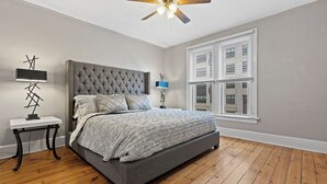 A bedroom with a tufted headboard bed, a bedside table with a lamp, a window with blinds, and a ceiling fan. - Stunning Townhouse in an Amazing Location Near Fiserv, Baird Center, Lakefront (Milwaukee)