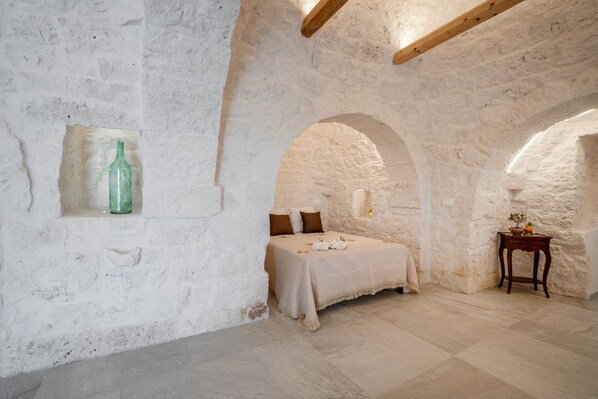 Une chambre rustique aux murs de pierre, avec un lit, une petite table et une bouteille fixée au mur.