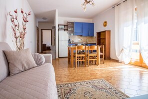 A living room with a sofa, a dining table with chairs, a kitchen area with blue cabinets, and a patterned rug.