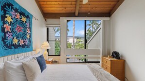 A bedroom with a bed, a nightstand, a lamp, a dresser, and a large window with a view of palm trees and a building.