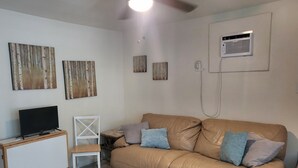 A living room with a tan leather sofa, a white chair, a small white cabinet, and a TV on a wooden stand. There are three wall-mounted artworks and a ceiling fan.