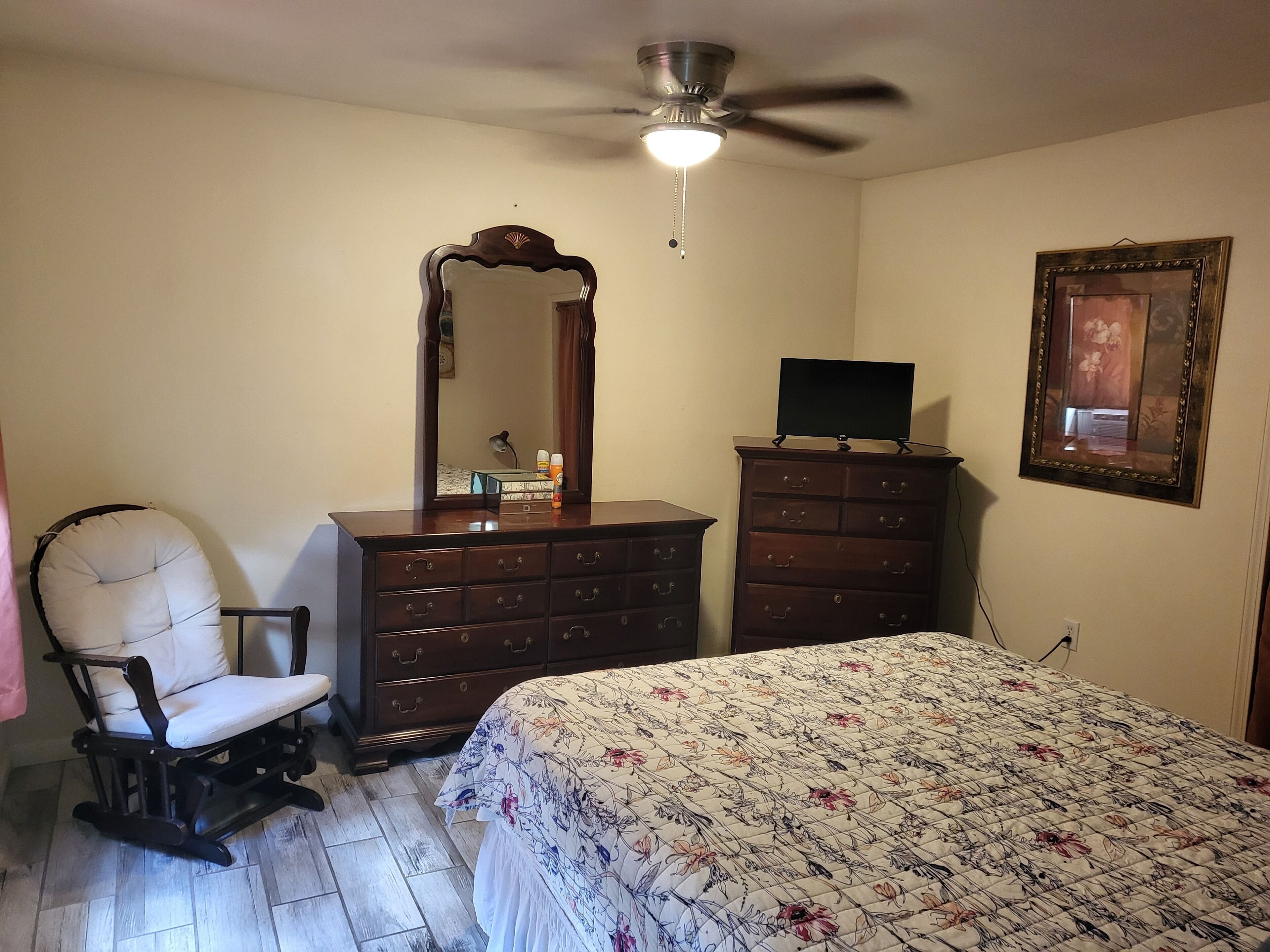 A bedroom with a bed, a dresser, a chair, a ceiling fan, and a mirror.