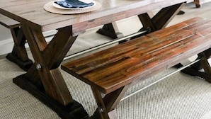 Une table à manger en bois, agrémentée de sets de table tressés et de serviettes bleues, un luminaire suspendu en fibres tressées et un vase avec des fleurs blanches.