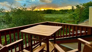 Une terrasse en bois avec une table et des chaises, donnant sur un champ verdoyant et des arbres sous un ciel nuageux.
