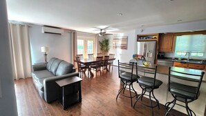 Ein modernes Wohnzimmer mit Sofa, Esstisch und Stühlen, eine Küche mit Kühlschrank und Spüle sowie ein Deckenventilator.