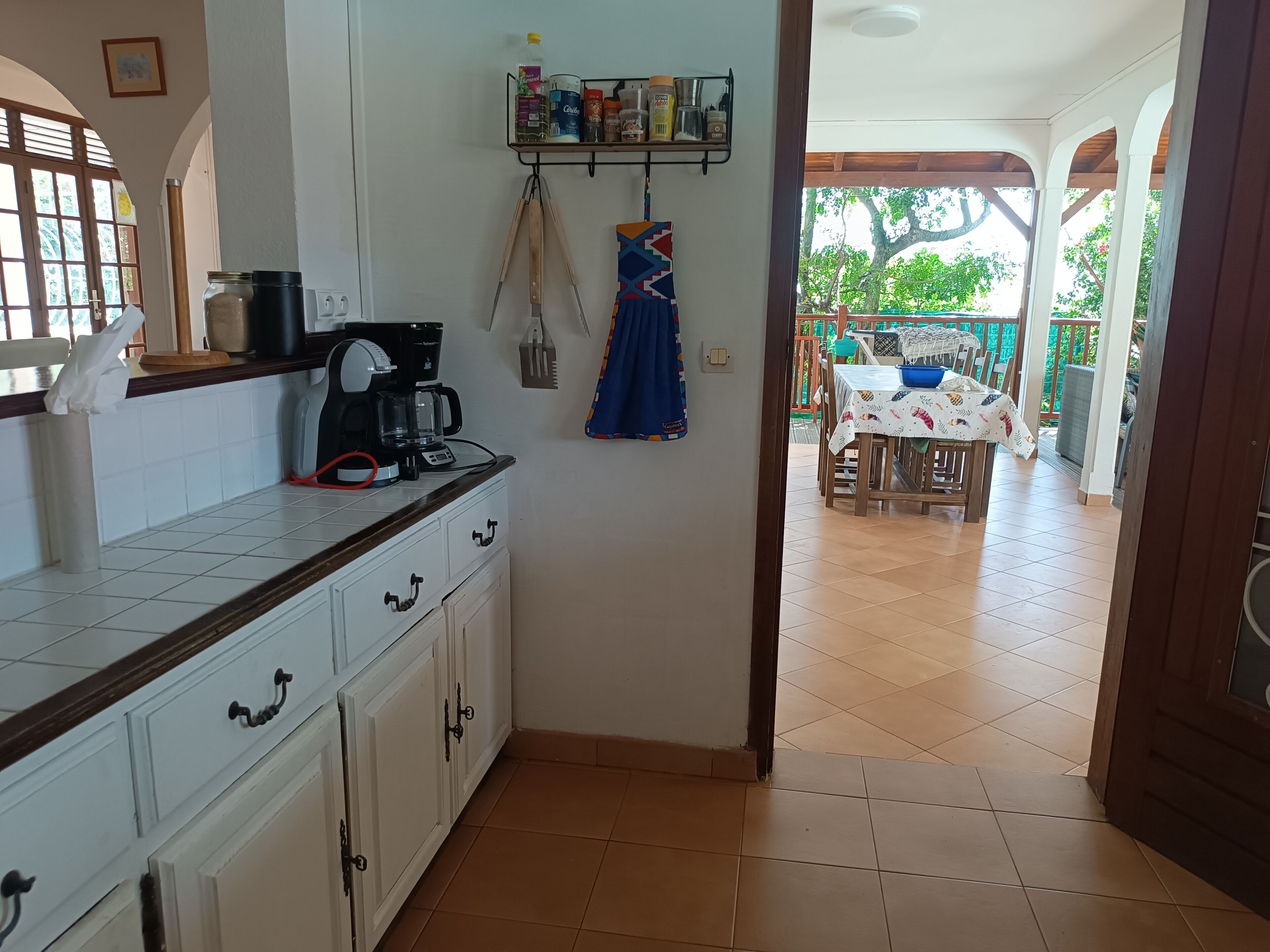 A kitchen with a coffee maker, jars, and a hanging towel. There is a dining area with a table and chairs outside.