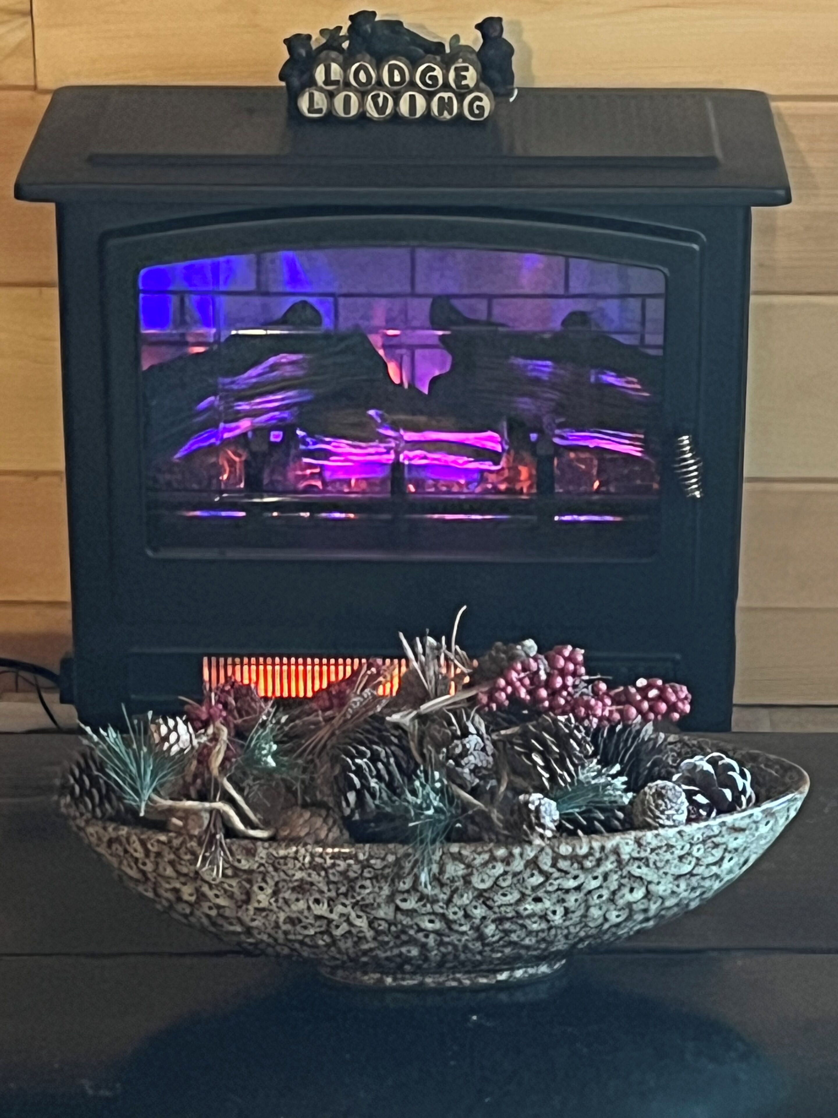 A wood-burning stove with a decorative sign above it and a bowl of pinecones and branches in front.