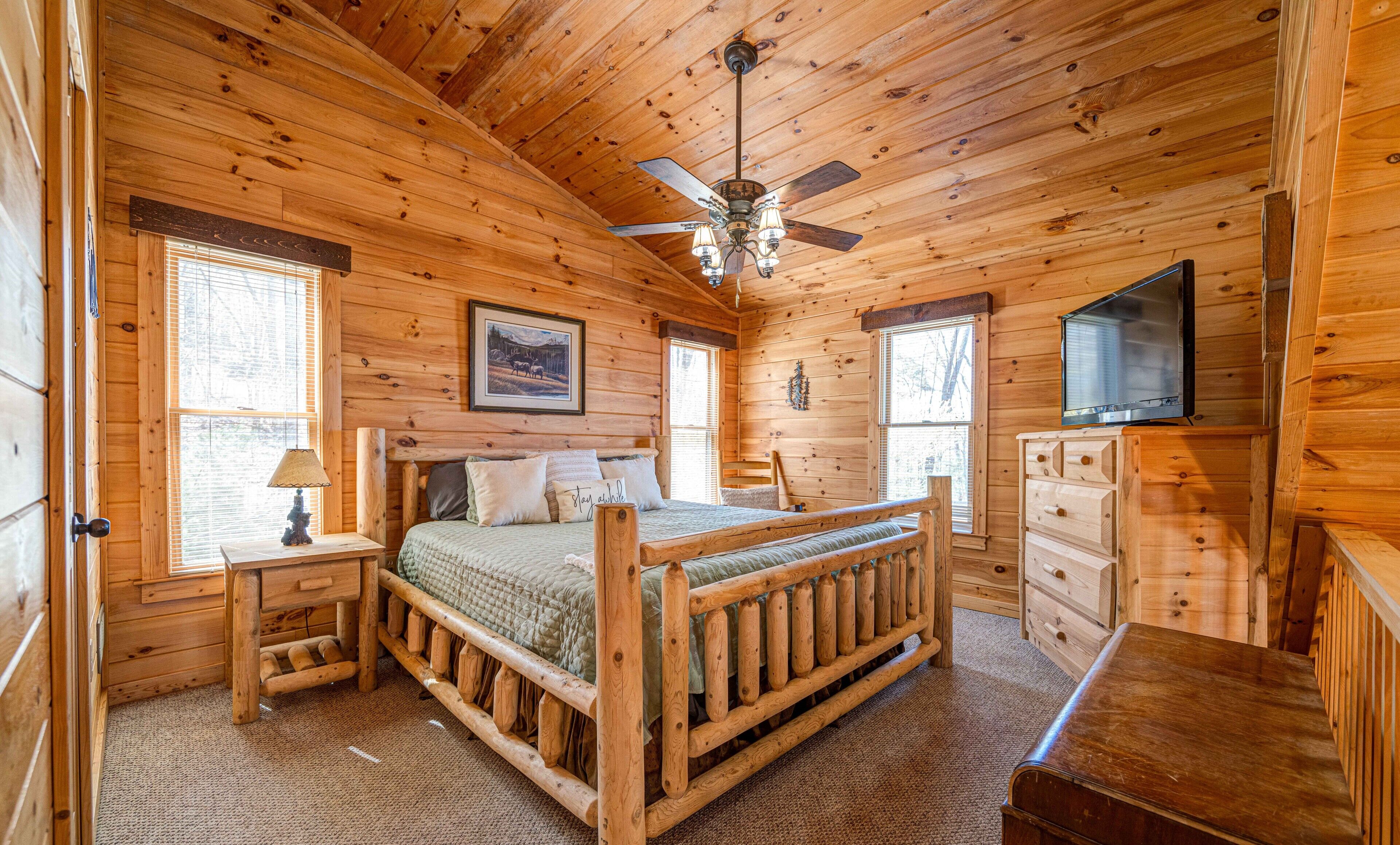 A wooden cabin bedroom with a bed, dresser, TV, and ceiling fan.