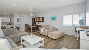 Un salon moderne avec un canapé, un pouf et une table basse. On y trouve également un coin repas avec une table et des chaises. La pièce est dotée d’une grande fenêtre avec des stores et d’un ventilateur de plafond.