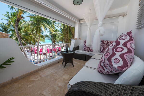 Un balcon avec un canapé blanc, une petite table et un auvent blanc. On aperçoit des palmiers et une plage à l’arrière-plan.