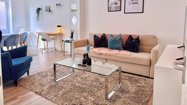 Un salon moderne avec un canapé beige, des fauteuils bleus, une table basse en verre et un tapis. On y trouve un bureau avec une chaise, une plante suspendue et des œuvres d’art murales.