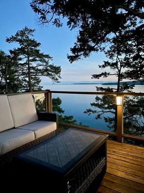 A deck with a sofa and a table, overlooking a body of water and distant land.