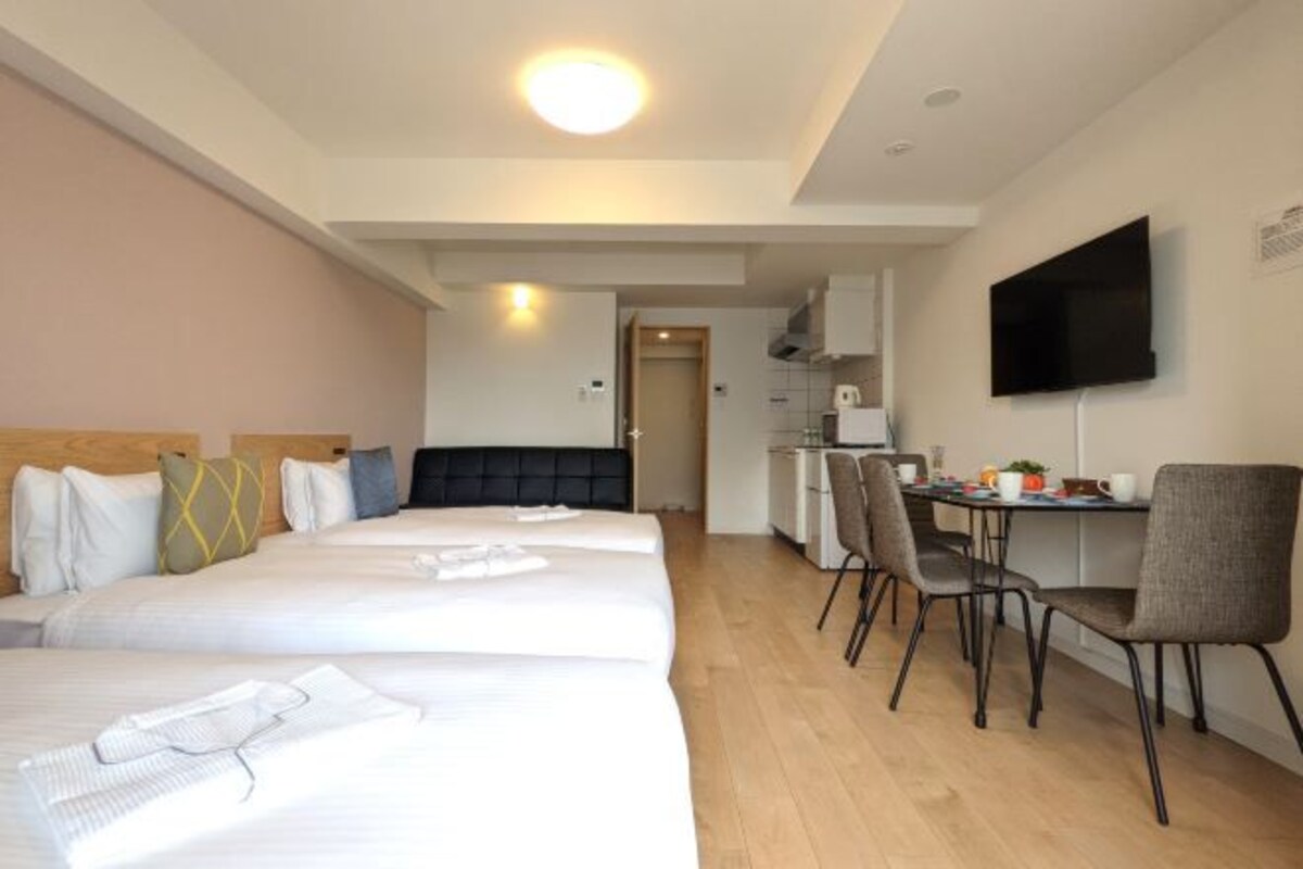 A hotel room with three beds, a desk with two chairs, a television, and a kitchenette.