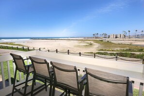 A beach view from a balcony with chairs, a chain-link fence, and a distant shoreline with buildings and palm trees.