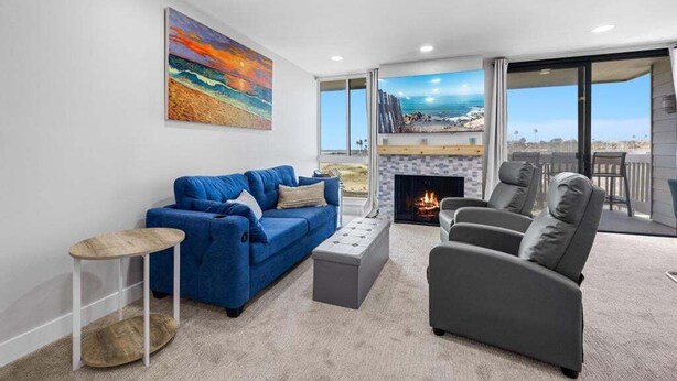 A modern living room with a blue sofa, grey armchair, and a small round table. A fireplace is visible, and there is a large painting of a beach scene on the wall.