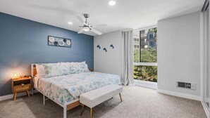 A bedroom with a bed, bedside table, bench, ceiling fan, artwork, and a large window with a view.