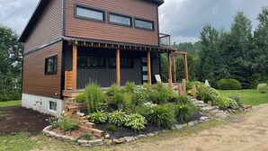 A modern two-story house with a wooden exterior, a balcony, and a garden with various plants and flowers. - Oasis thermal -  Mauricie- N.E. Chic Shack (Saint-Mathieu-du-Parc)