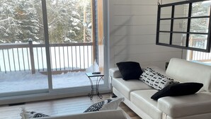 A modern living room with a white sofa, a black and white patterned pillow, and a black throw pillow. There is a window with a view of a snowy outdoor area. - Oasis thermal -  Mauricie- N.E. Chic Shack (Saint-Mathieu-du-Parc)
