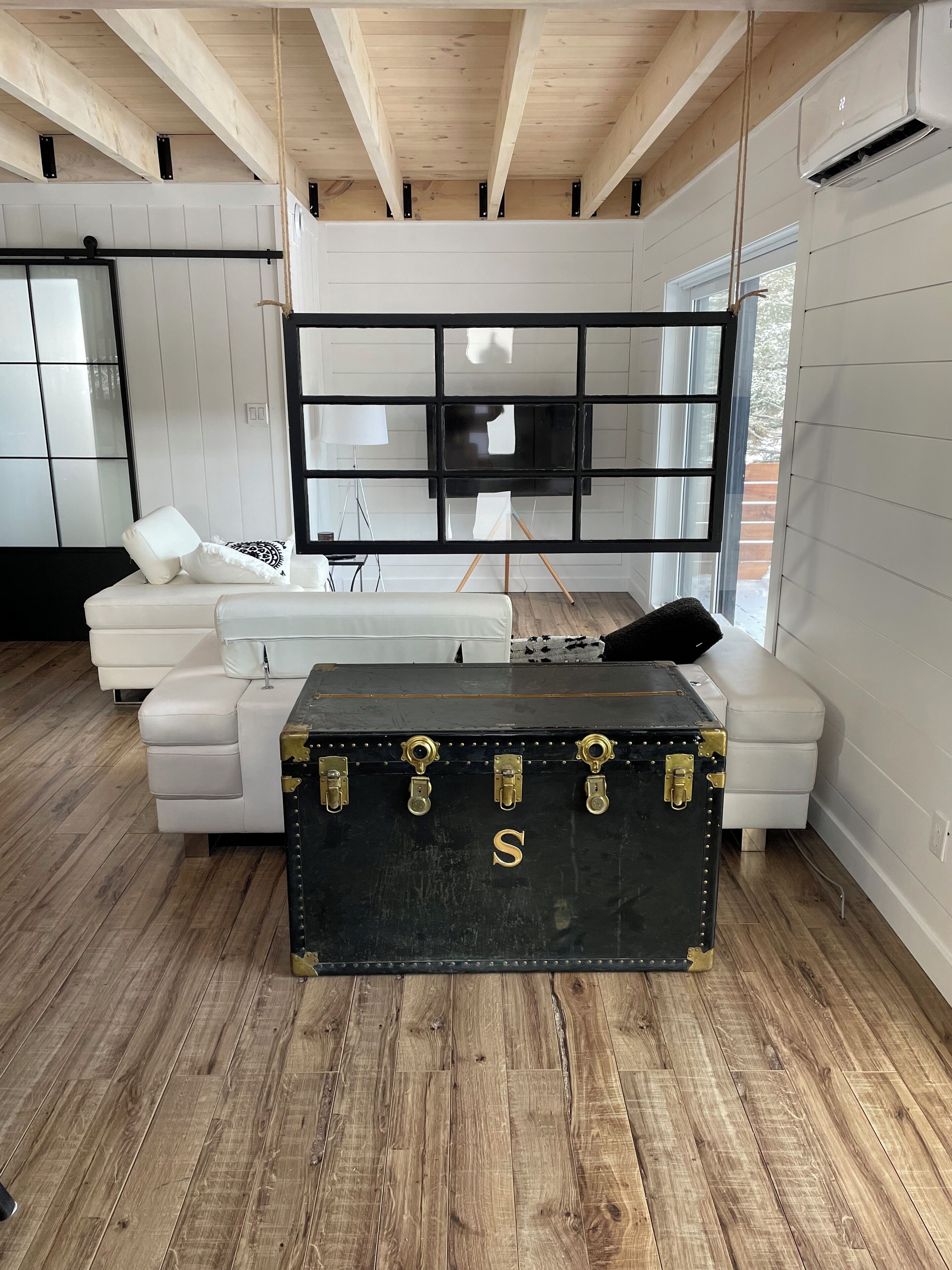 A modern living room with a white sofa, a black trunk with gold locks, and a large window.