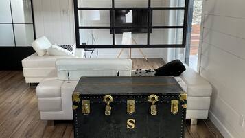 A modern living room with a white sofa, a black trunk with gold locks, and a large window.