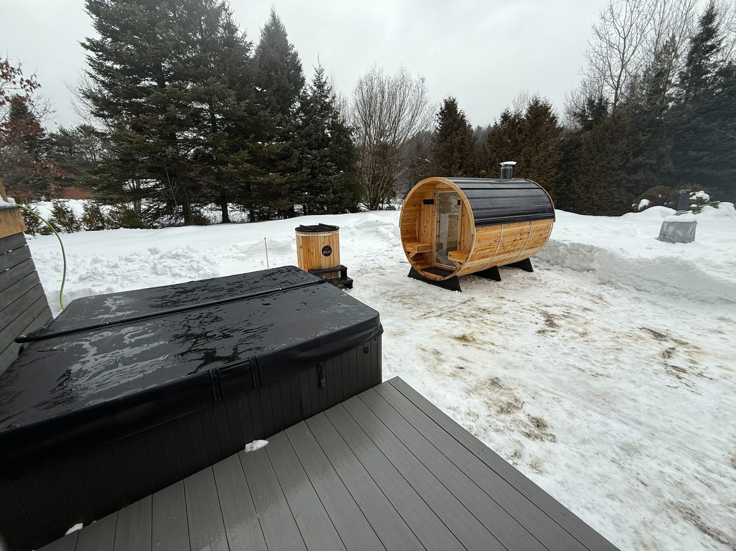 A wooden sauna structure and a hot tub on a snow-covered deck.