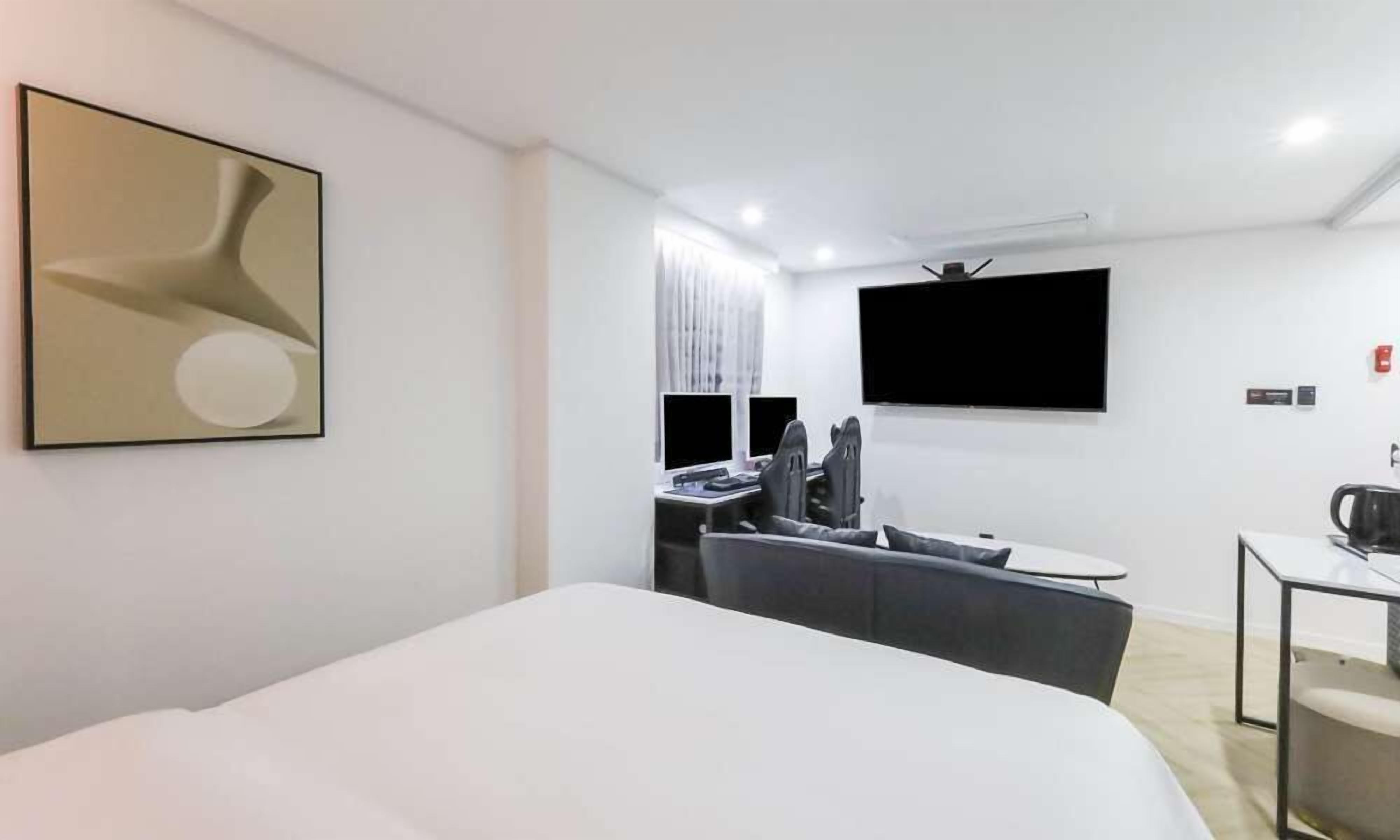 A modern hotel room with a large bed, a desk with two monitors, a sofa, and a flat-screen TV mounted on the wall.