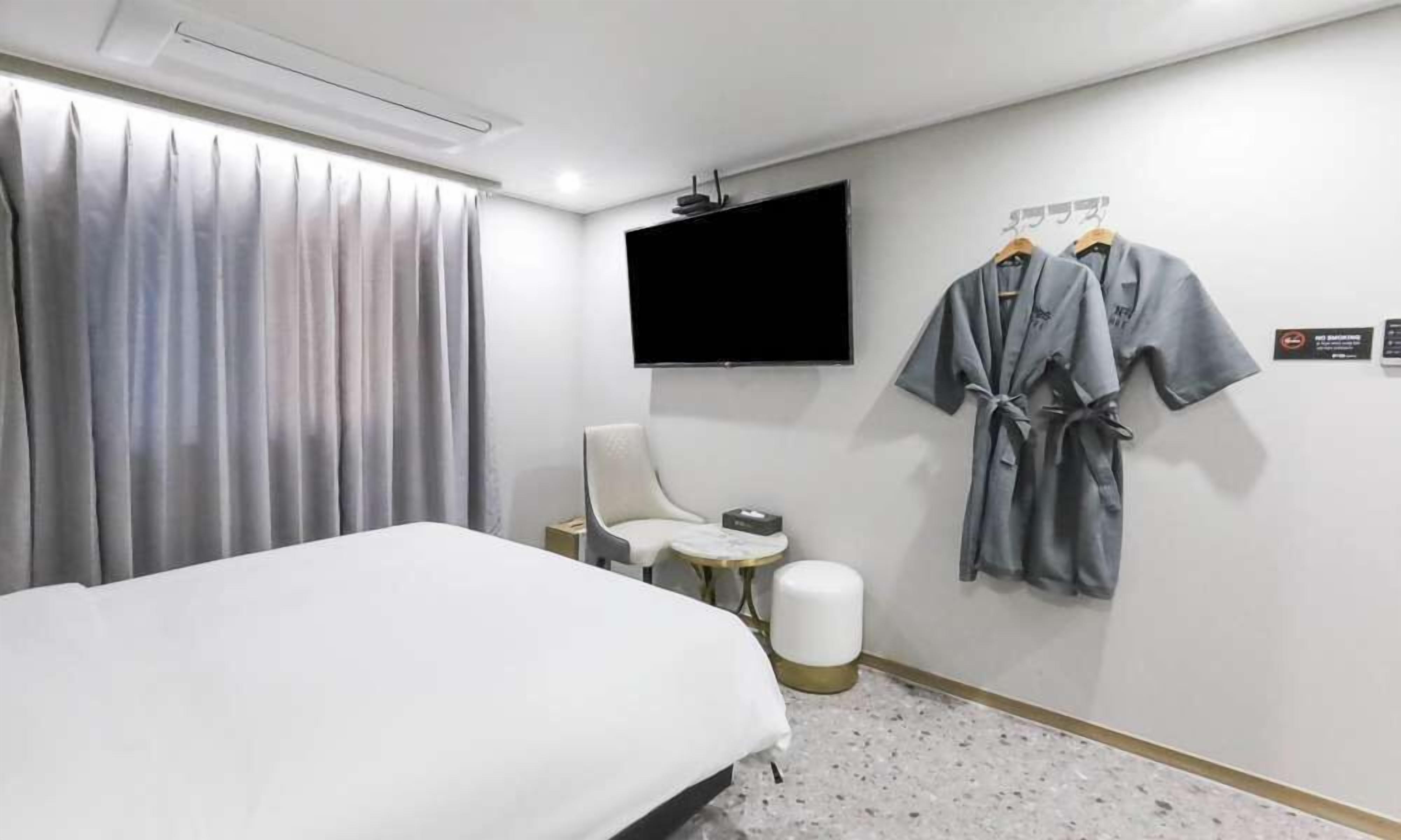 A hotel room with a bed, a chair, a small table, a TV mounted on the wall, and two robes hanging on a rack.