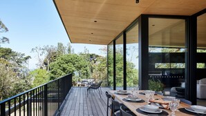 Et moderne udendørs spiseområde med træterrasse, metalgelænder og glasvægge, der giver udsigt til de omkringliggende træer og landskab.