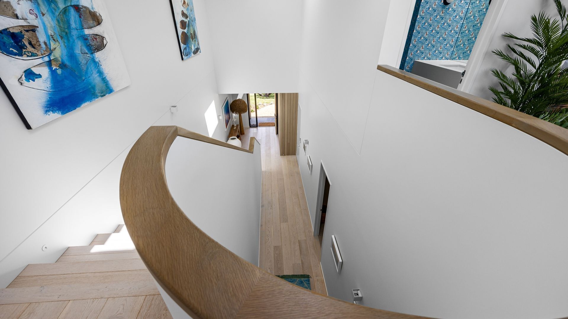 Un intérieur moderne avec un escalier en bois aux lignes courbes, un couloir agrémenté d’œuvres d’art et une vue sur une pièce dont le mur est orné de motifs.