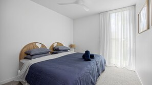 Een slaapkamer met een bed, twee hoofdeinden, een bedlampje, een raam met dunne gordijnen en een plafondventilator.