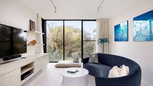 A modern living room with a flat-screen TV, a dark sofa, a round coffee table, and a large window offering a view of trees and a clear sky.
