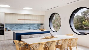 A modern kitchen with a central island, a dining area with wicker chairs, and large windows offering a view of trees.