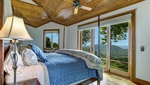 Une chambre à coucher avec un lit, une lampe et une vue sur un paysage montagneux, visible à travers de grandes fenêtres.