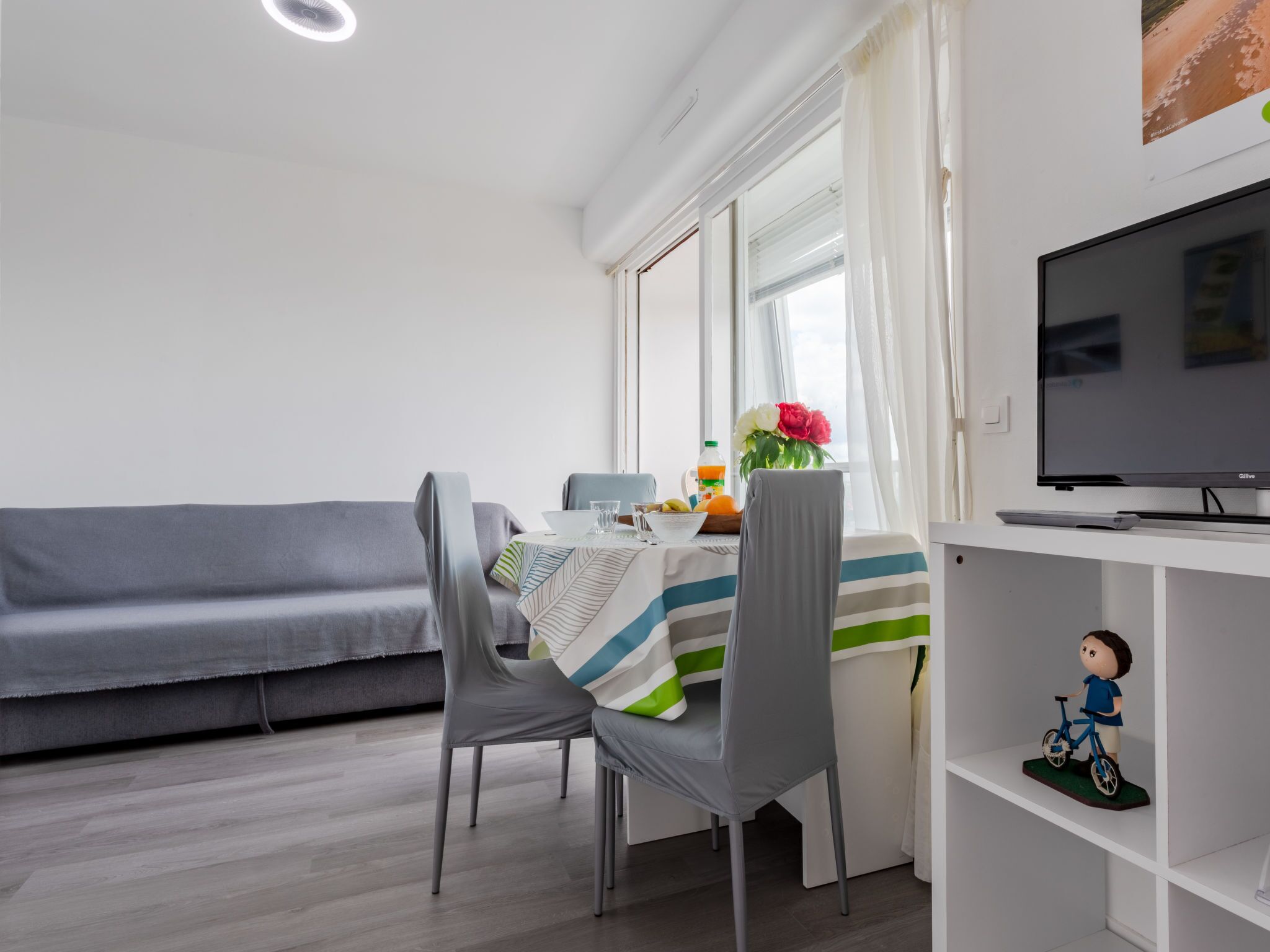 A modern living room with a grey sofa, a dining table with chairs, a TV on a white shelf, and a window with curtains.
