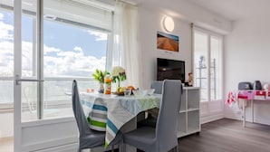A modern dining area with a table set for two, a TV mounted on the wall, and a view of the sky through large windows.